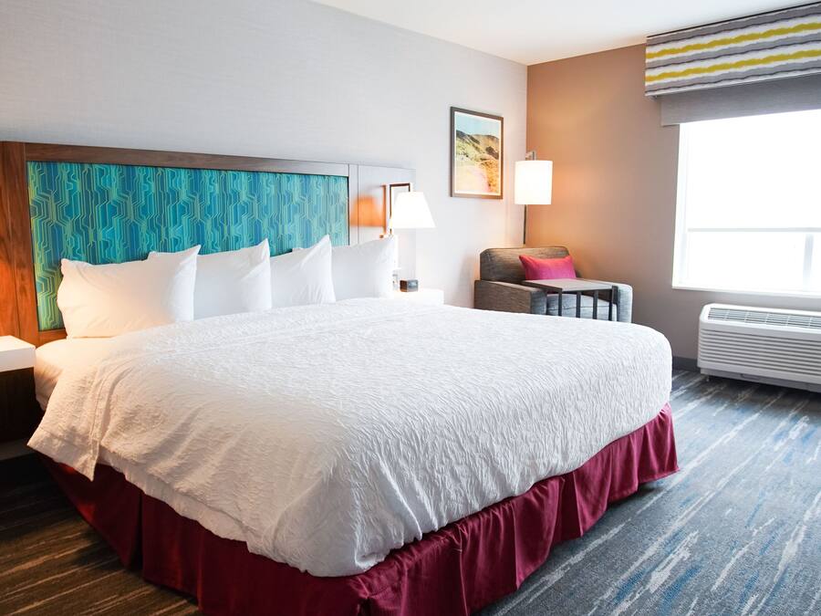 Une chambre d’hôtel avec un grand lit, deux lampes de chevet, un téléphone fixé au mur, une petite table avec une chaise et une fenêtre avec un climatiseur.