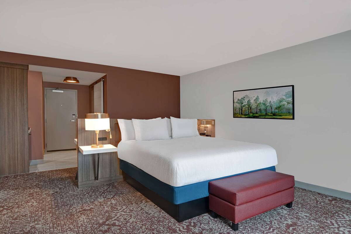 A hotel room with a large bed, a bedside table with a lamp, a red bench, a painting on the wall, and a door leading to another room.