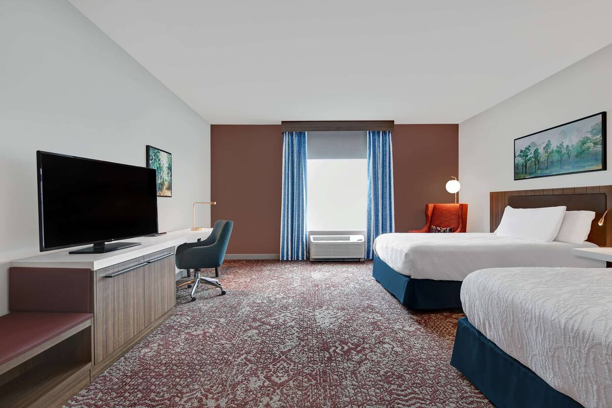 A hotel room with a flat-screen TV, a desk, a chair, two beds, a large window with curtains, a painting on the wall, and a lamp.