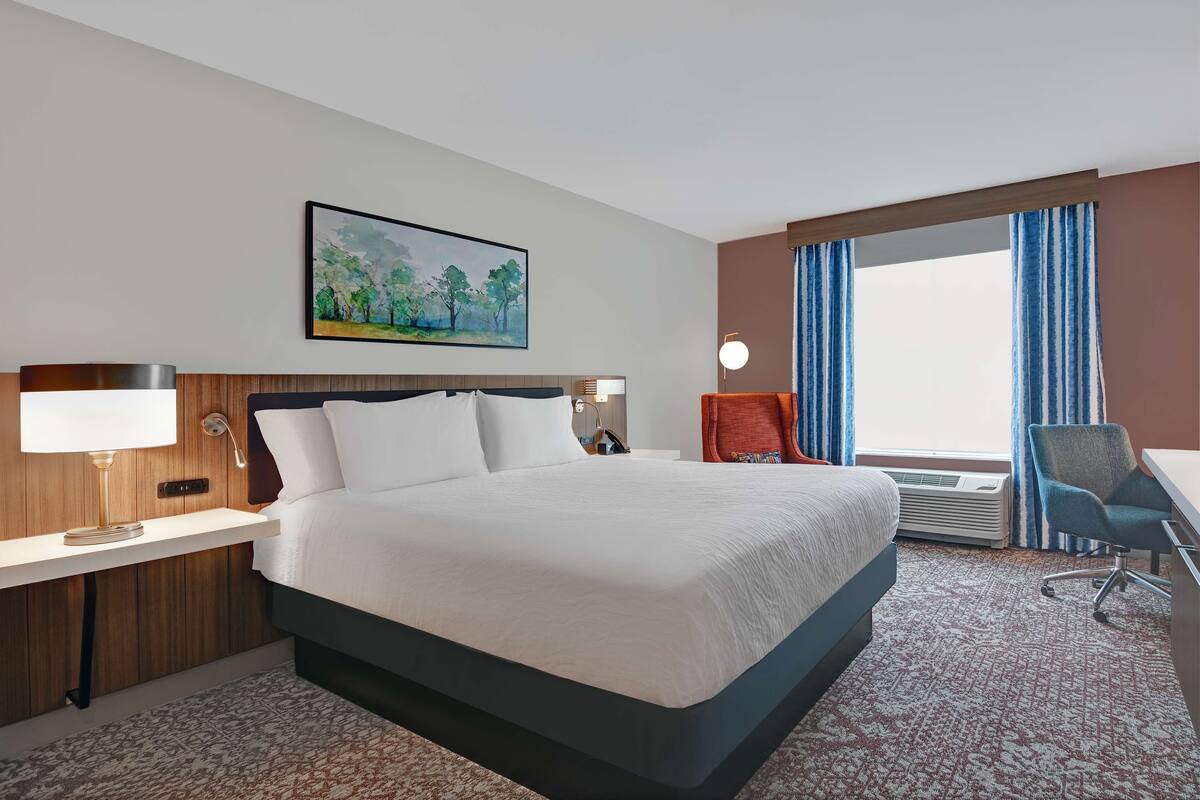 A hotel room with a large bed, a desk, a chair, a painting on the wall, and a window with curtains.