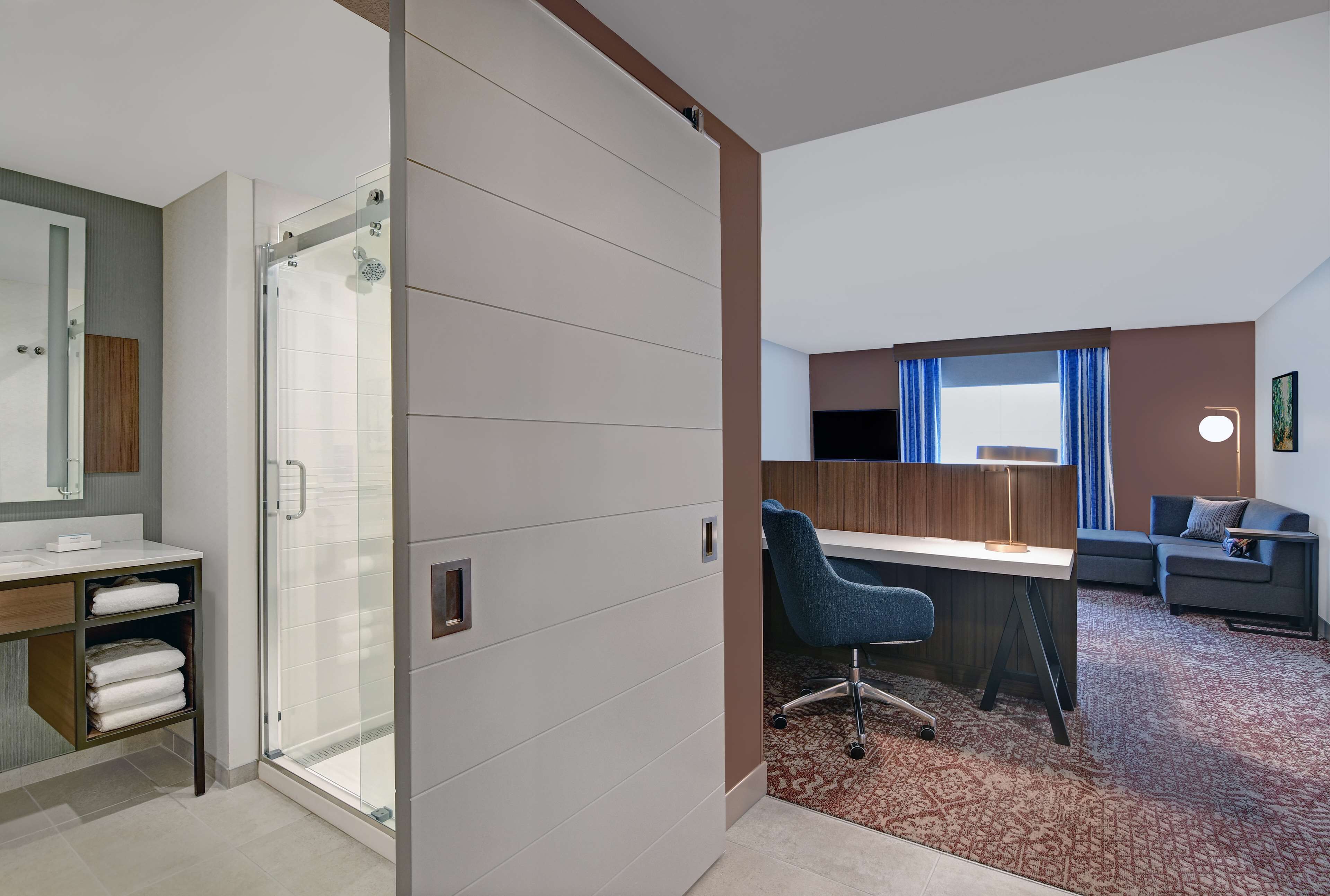 A hotel room with a shower, a desk, a chair, a sofa, and a patterned carpet.