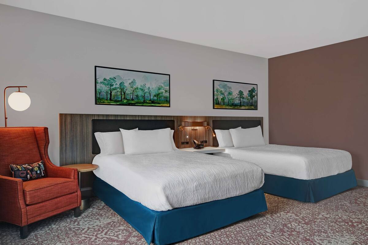 A hotel room with a double bed, a side table, a chair, and two framed pictures on the wall.