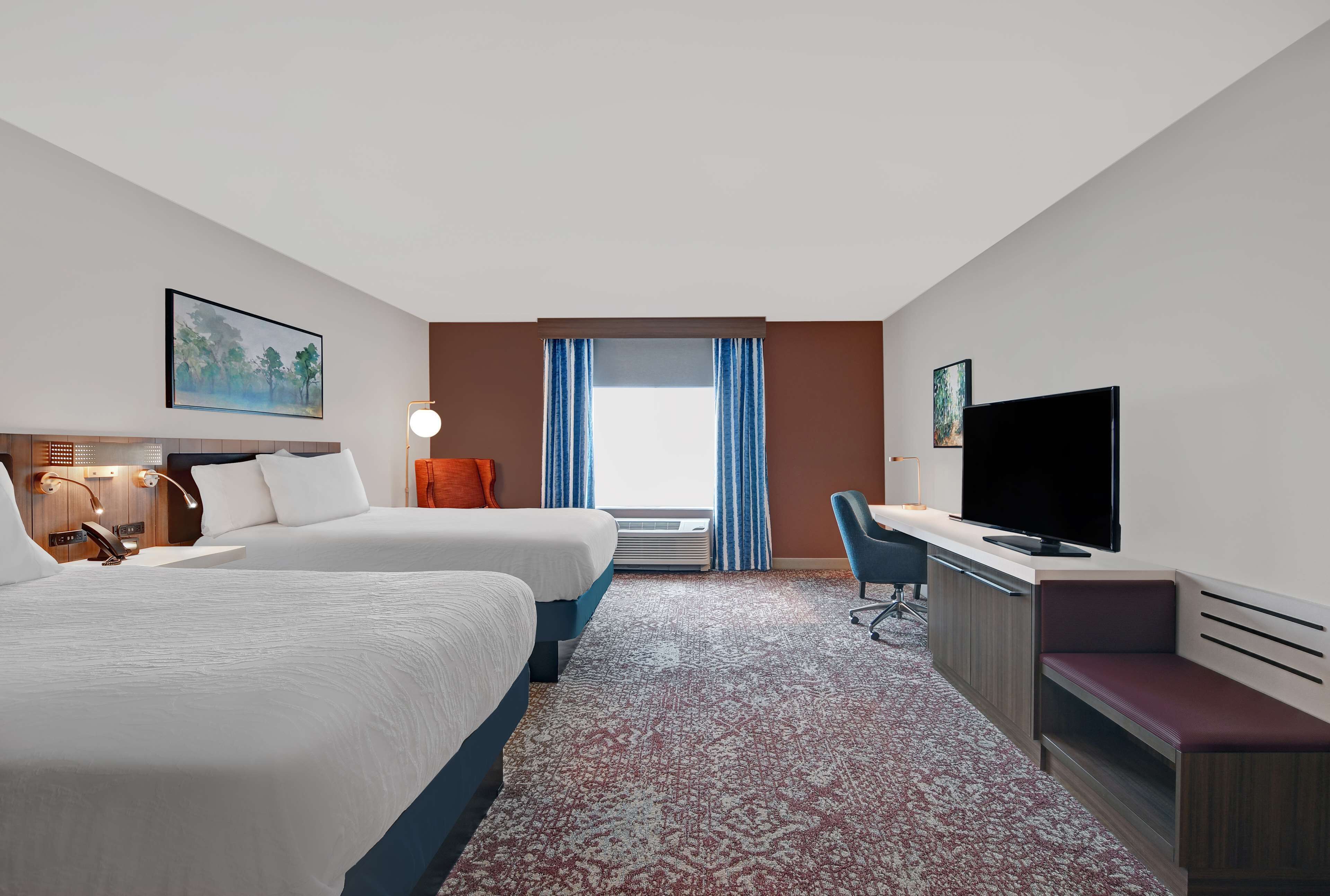 A hotel room with two beds, a desk with a TV, a desk lamp, a chair, and a window with curtains.