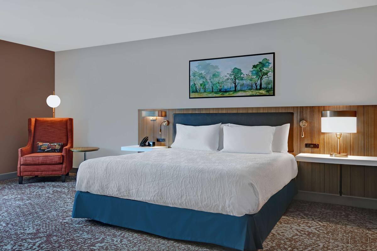 A hotel room with a large bed, a red armchair, a bedside table with a lamp, and a painting on the wall.