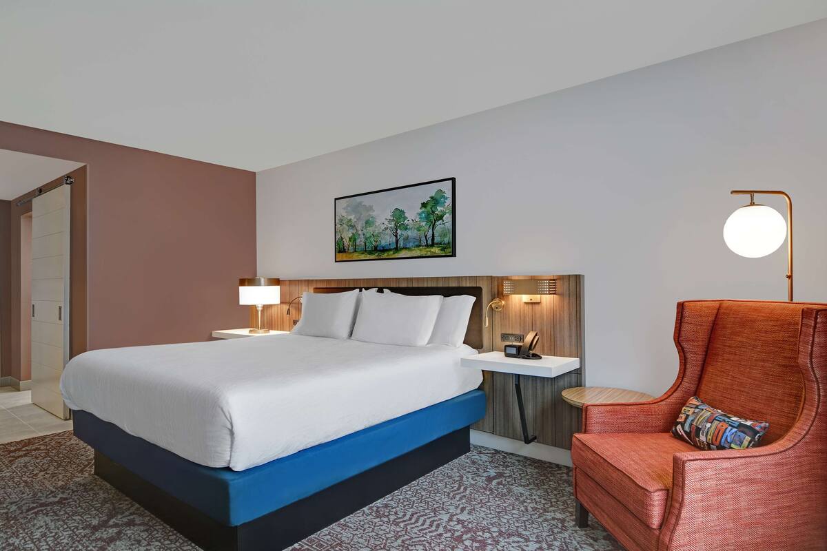 A hotel room with a large bed, a side table with a lamp, a chair with a colorful cushion, and a painting on the wall.