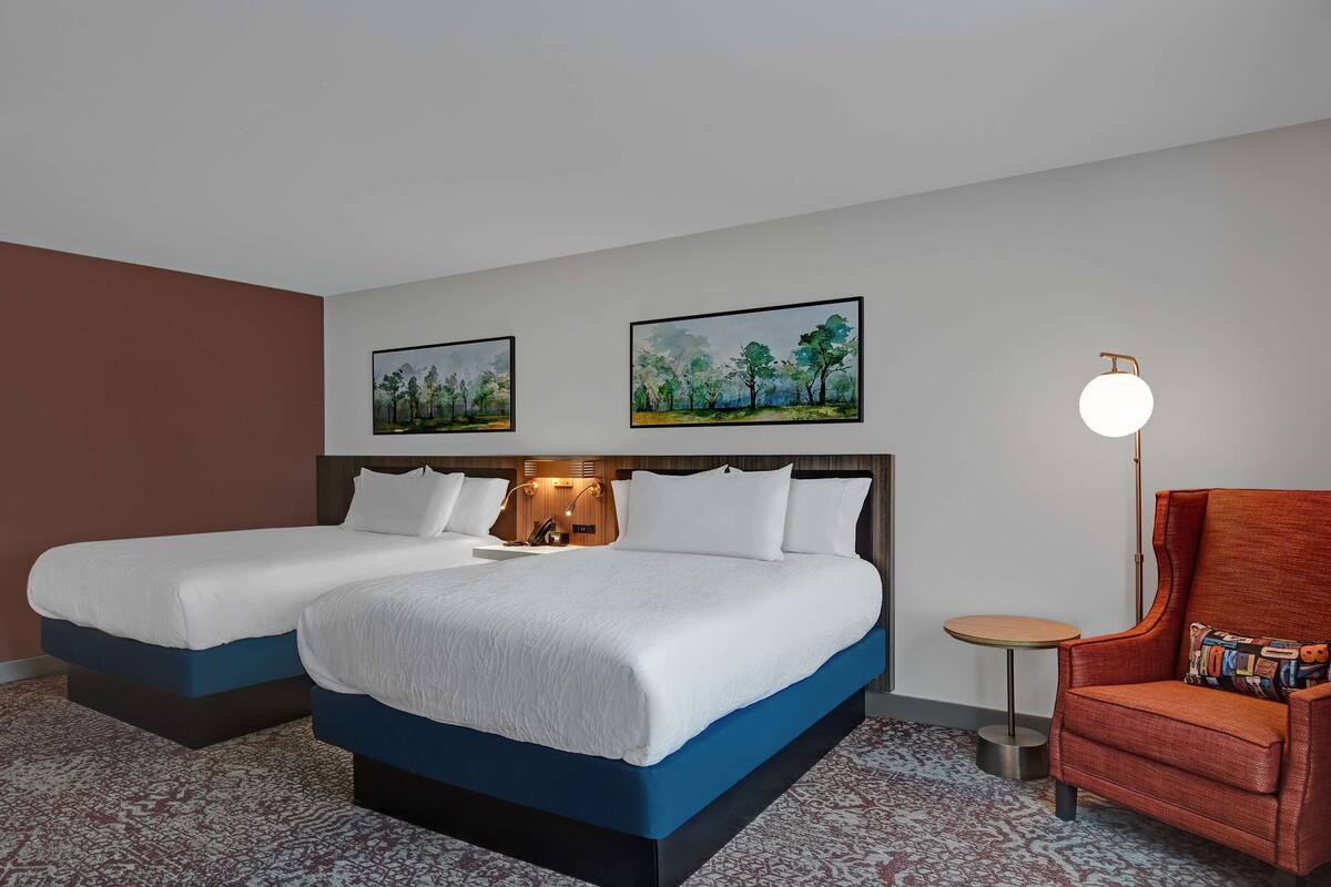 A hotel room with two beds, a small round table, a chair, and two framed pictures on the wall.