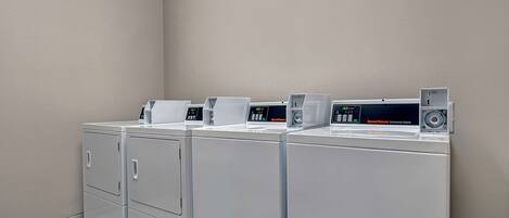 A row of white commercial washers with control panels, in a clean, tiled laundry room with beige walls.