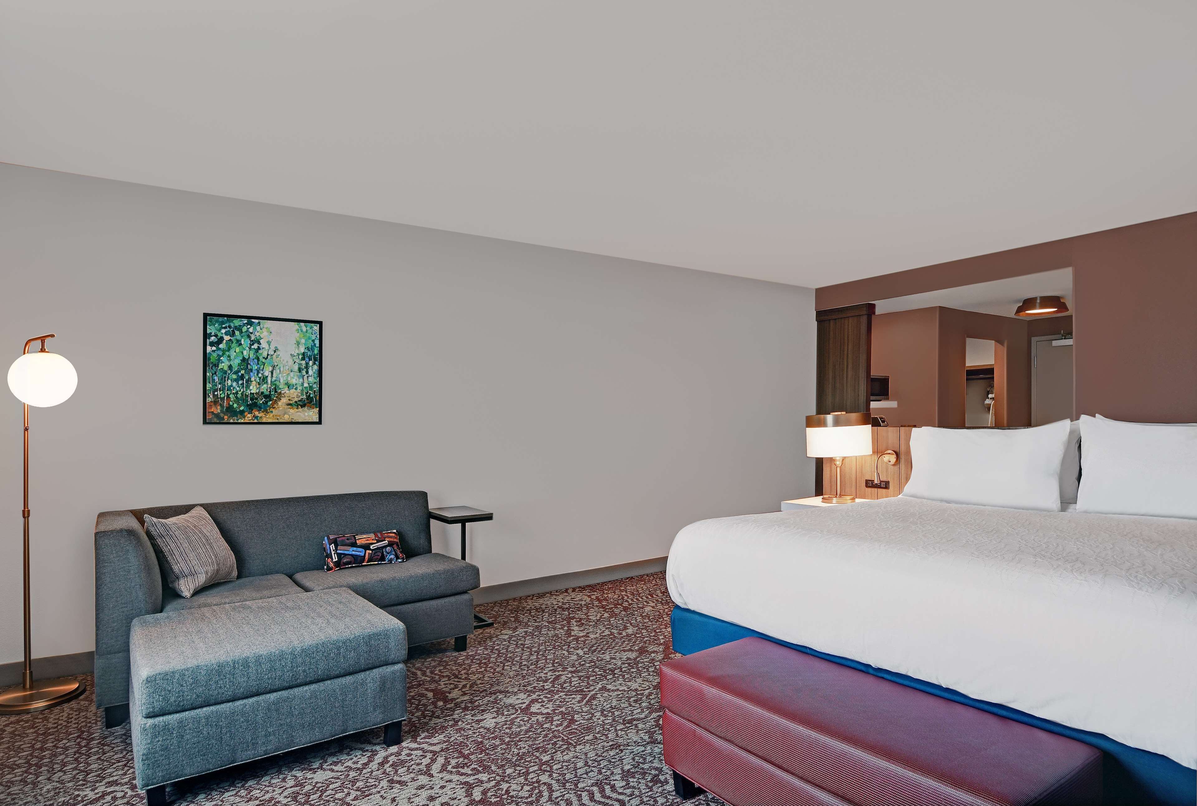 A hotel room with a bed, a sofa, a small table, a lamp, and a painting on the wall.