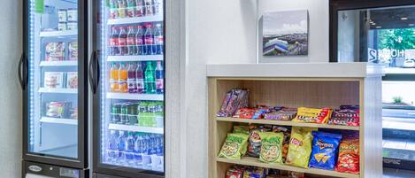 Two self-service beverage dispensers stocked with various drinks, a snack shelf with chips and packaged foods, and a framed picture on the wall.