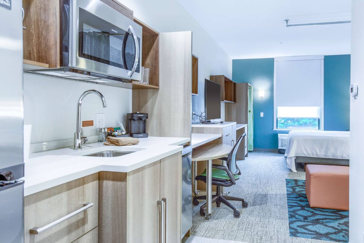 A compact hotel room with a kitchenette, a bed, a desk, and a window.
