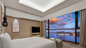 Habitación de hotel con una cama grande, un televisor de pantalla plana, una obra de arte en la pared y vista a un cuerpo de agua con edificios tradicionales.
