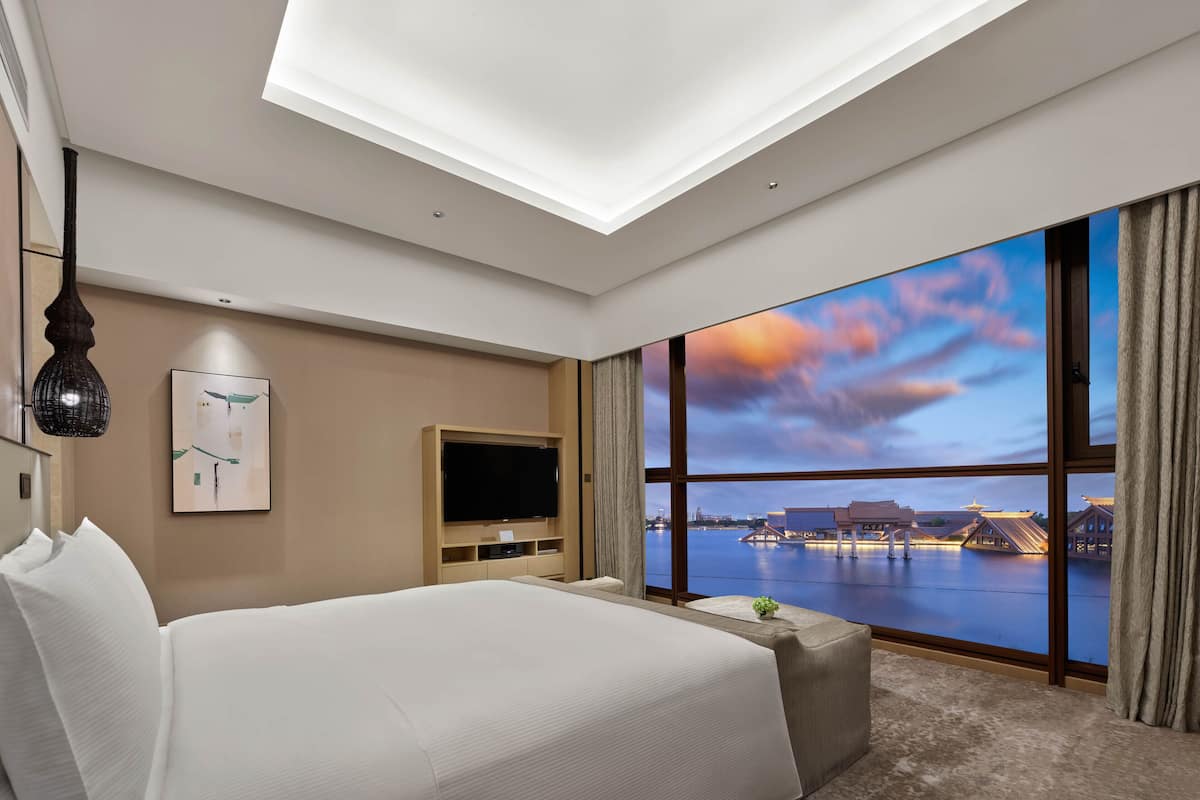 A hotel room with a large bed, a flat-screen TV, a wall-mounted artwork, and a view of a body of water with traditional buildings.