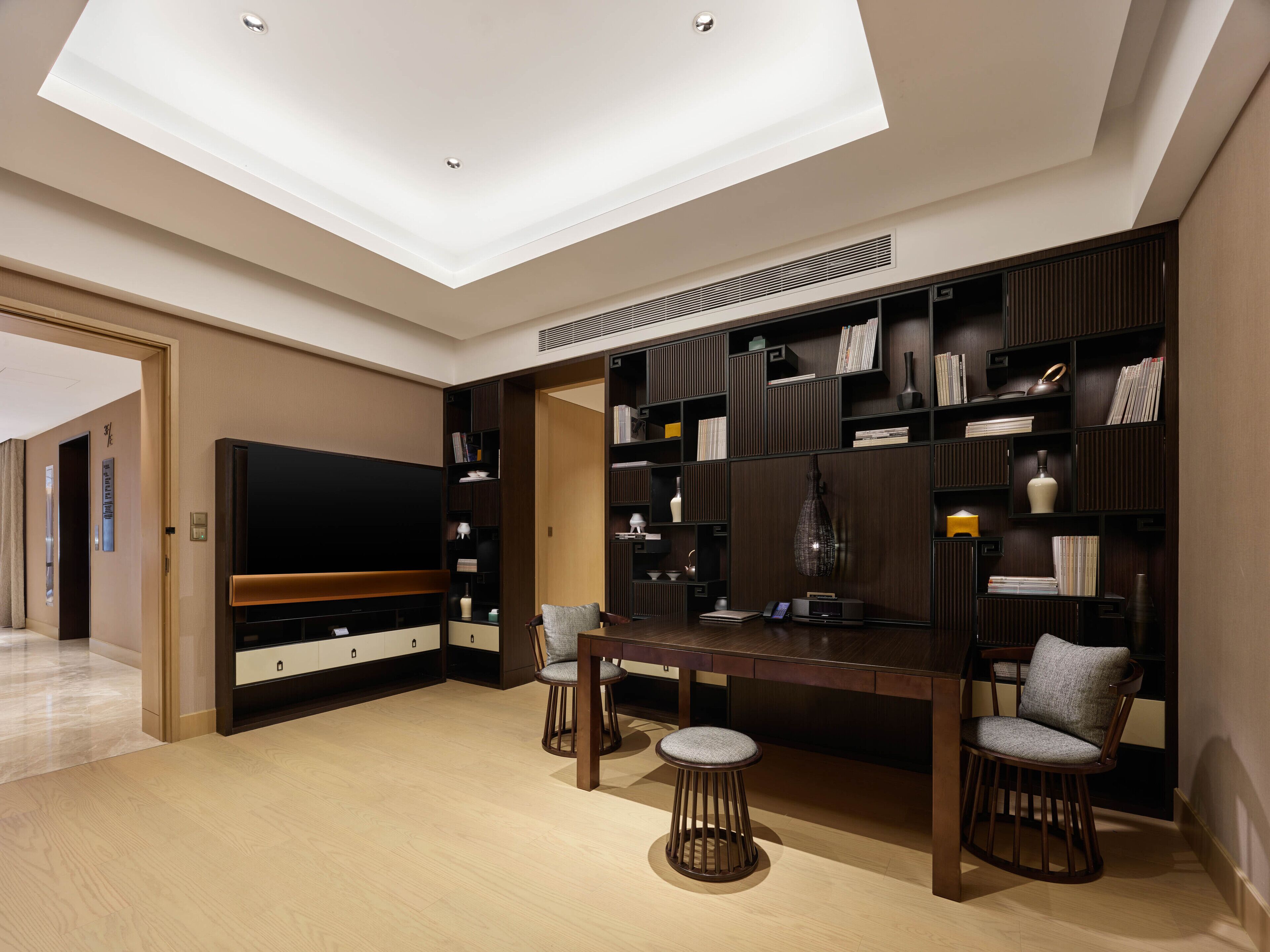A modern living room with a flat-screen TV, dark wood shelving, a dining table, and two bar stools.