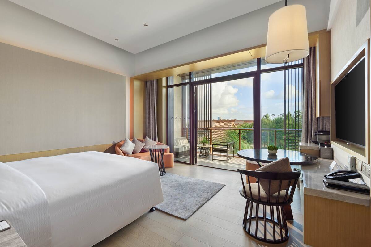 A modern hotel room with a large bed, a desk with a chair, a dining area with a table and chairs, and a balcony with a view of the city.