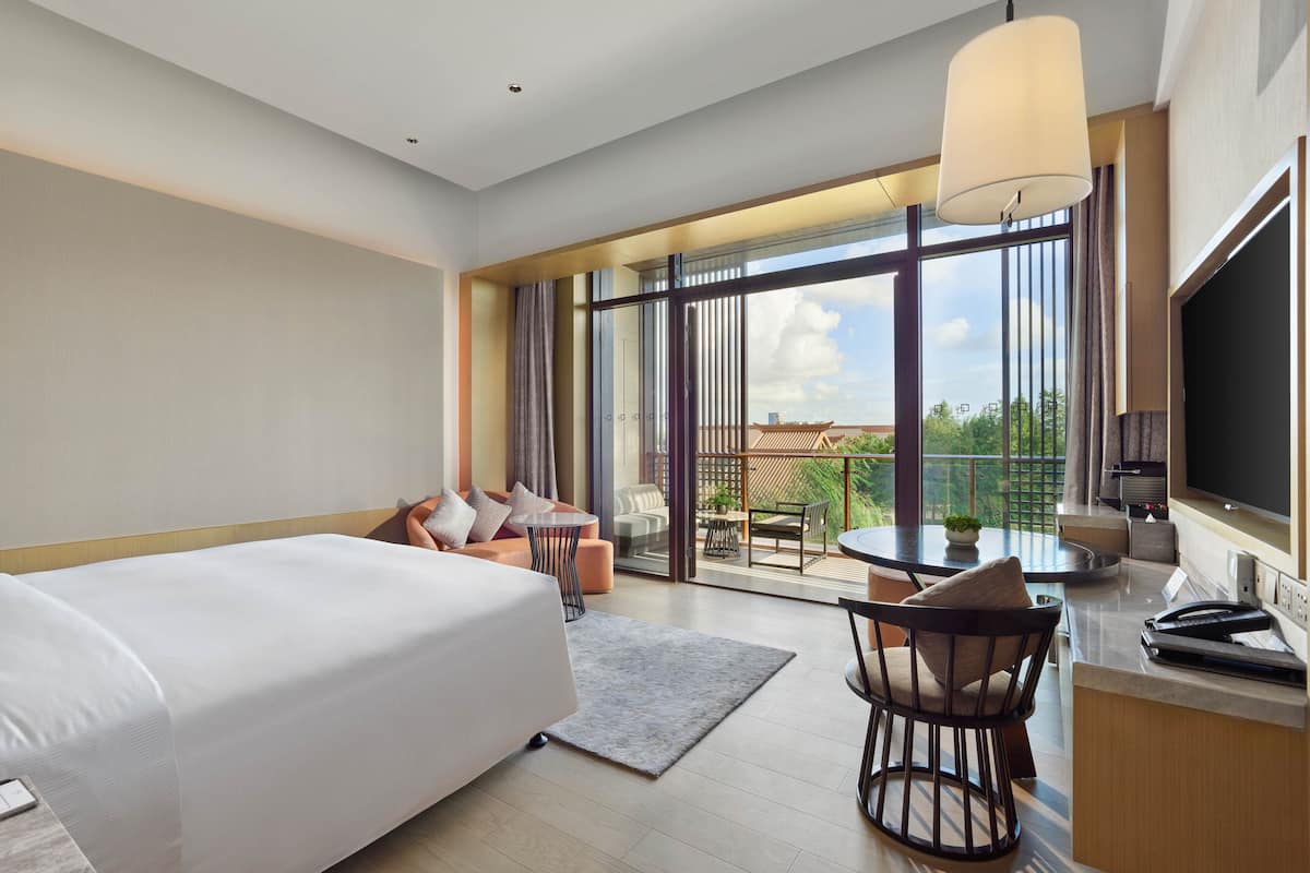 Une chambre d’hôtel moderne dotée d’un grand lit, d’un bureau avec une chaise, d’un coin repas avec une table et des chaises, et d’un balcon offrant une vue sur la ville.
