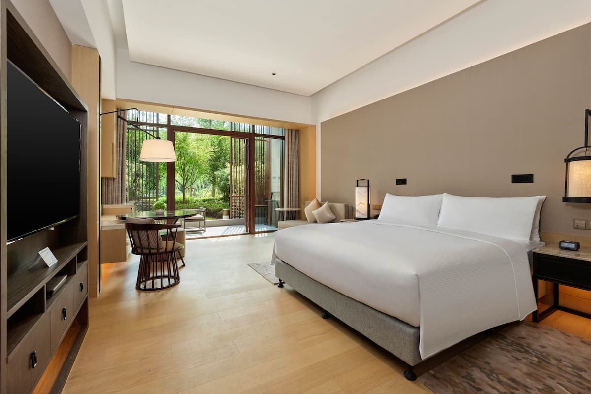 A modern hotel room with a large bed, a TV, a dining area with a table and chairs, and a view of greenery through large windows.