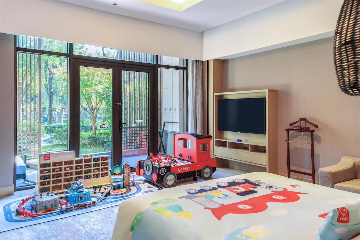 A child's playroom with a train set, a large window with a view of trees, and a television on the wall.