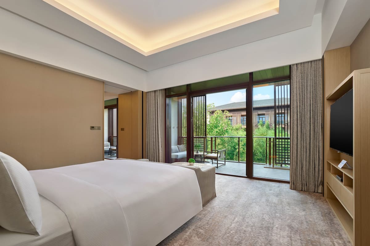 A modern hotel room with a large bed, a TV, and a balcony with a view of the building and greenery.