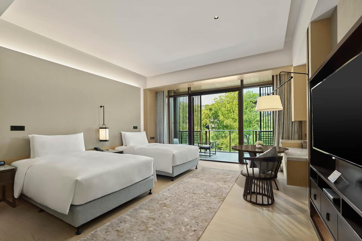 A modern hotel room with two beds, a desk with a chair, a flat-screen TV, and a balcony with a view of greenery.