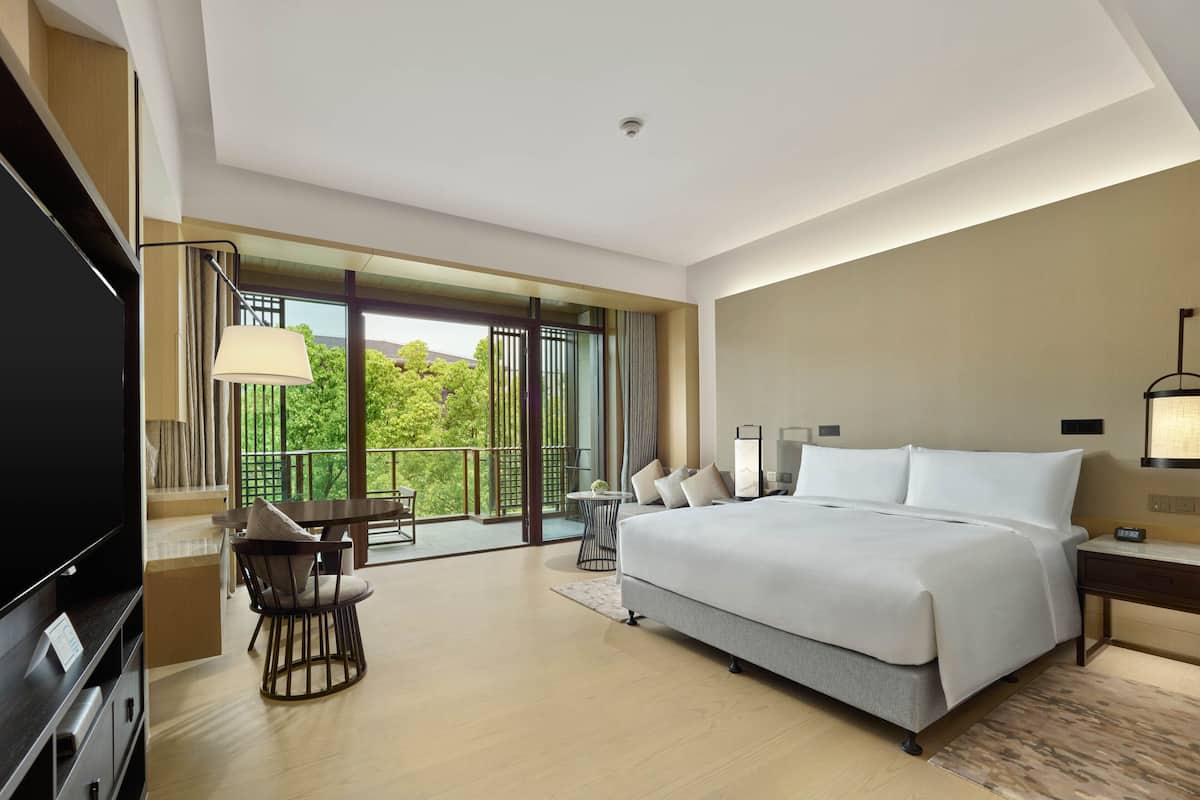 A modern hotel room with a large bed, a desk, a chair, and a balcony with a view of trees.