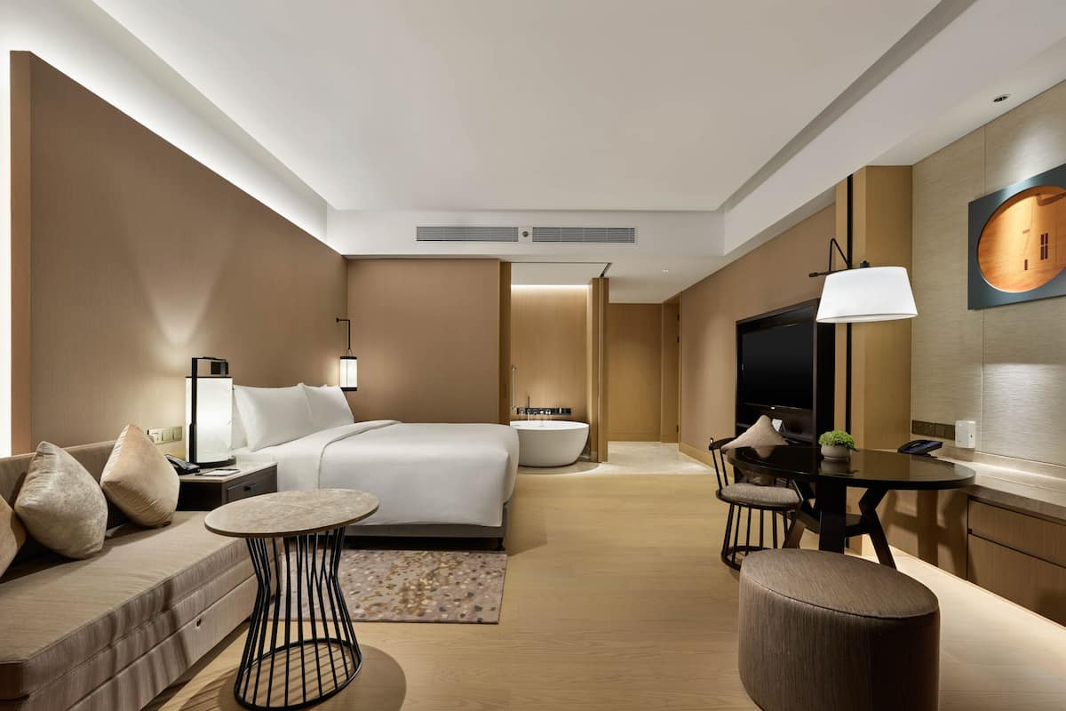 A modern hotel room with a bed, sofa, dining table, and a TV.