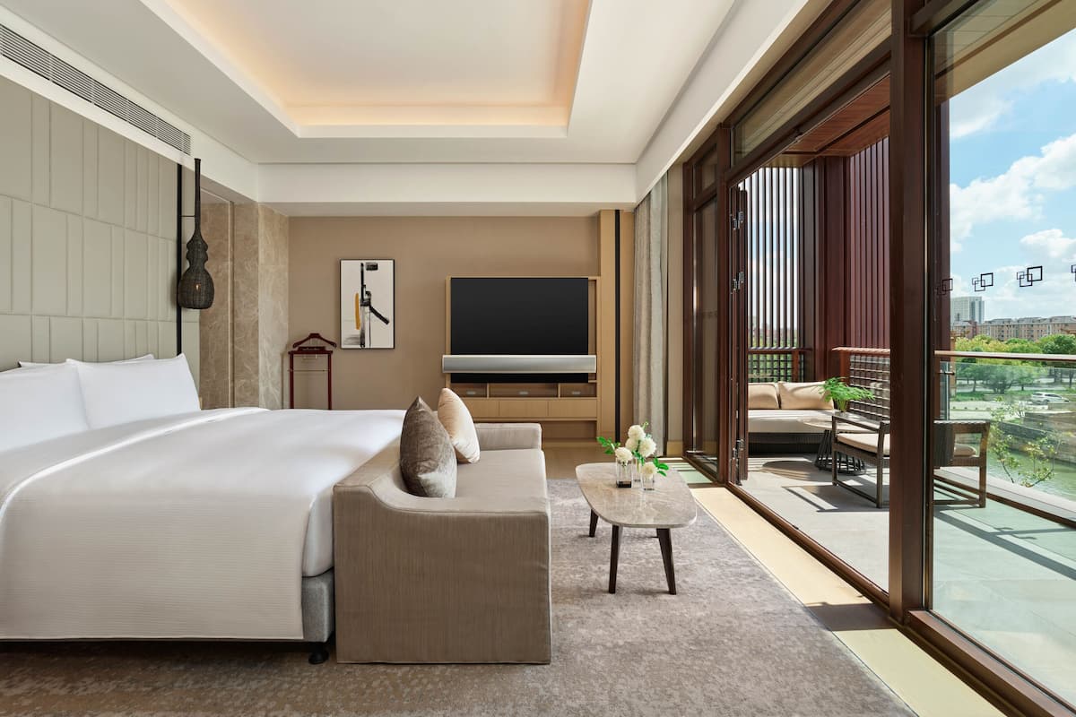 A modern hotel room with a large bed, a sofa, a TV, and a balcony with outdoor furniture.