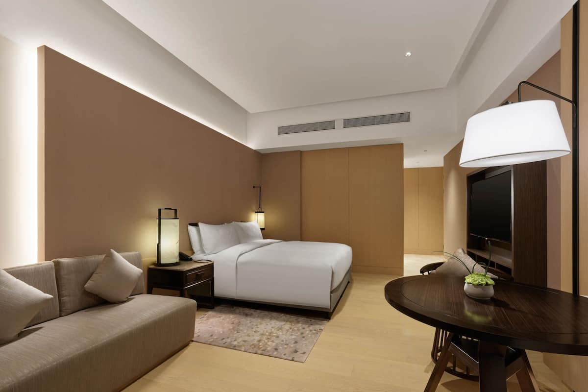 A modern hotel room with a bed, a sofa, a dining table, and a TV.