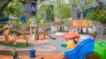 Un parque infantil con diversas estructuras de juego, incluyendo toboganes y zonas de escalada, rodeado de árboles y edificios.