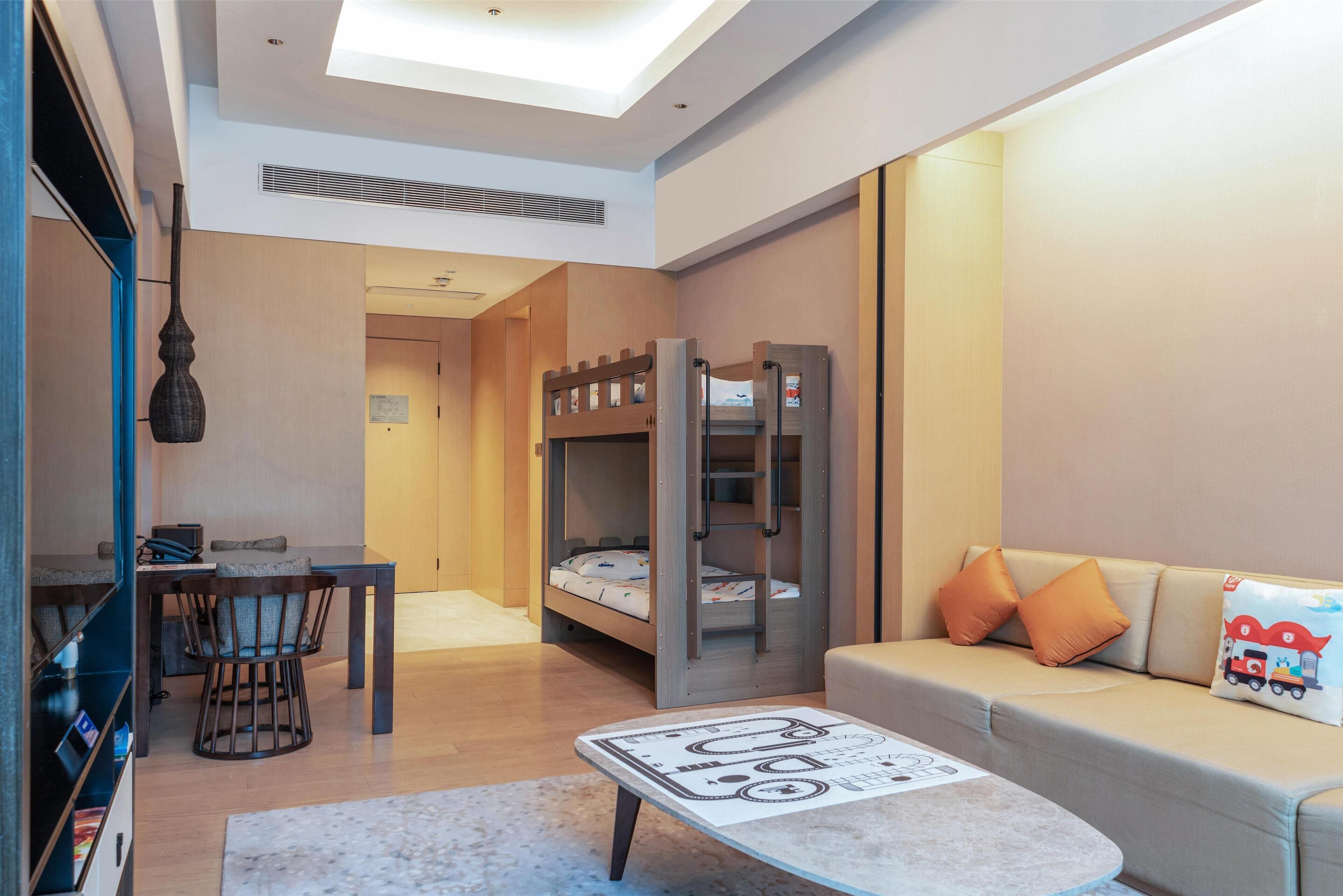 Una habitación de hotel moderna con una cama litera, un área de comedor con una mesa y sillas, un sofá con cojines y una pequeña mesa con un mantel individual.
