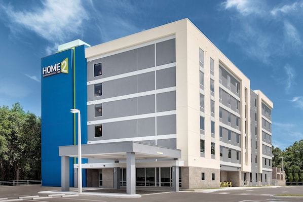 Ein modernes Hotelgebäude mit einer auffälligen blauen Schilder, auf denen "HOME 2 Suites & Hilton" steht. Es hat eine mehrstöckige Struktur mit großen Fenstern und einem überdachten Eingangsbereich.