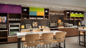 A modern cafe with a long communal table, multiple chairs, and a variety of food and drink options. - Home2 Suites by Hilton Tampa Westshore Airport, FL (Tampa)