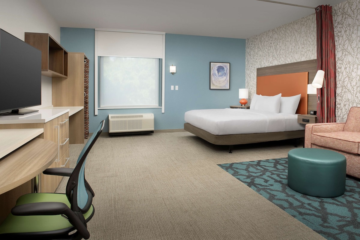 Ein Hotelzimmer mit einem Bett, einem Schreibtisch mit Stuhl, einem Fernseher, einem Fenster und einem gemusterten Teppich.