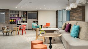 A modern hotel lobby with a sofa, ottomans, a round table, and a TV in the background. - Home2 Suites by Hilton Tampa Westshore Airport, FL (Tampa)