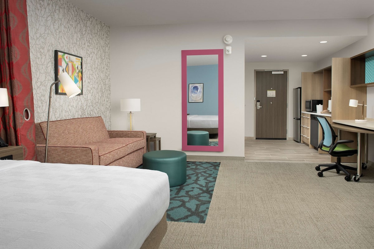Ein Hotelzimmer mit einem Bett, einem Sofa, einem Schreibtisch, einem Sessel, einer Lampe, einem Spiegel und einer Tür zu einem Badezimmer.
