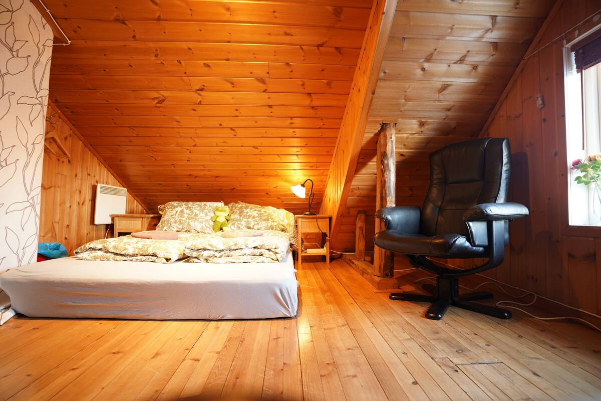 A cozy attic bedroom with a wooden ceiling, a bed with a white comforter, a black leather chair, and a nightstand with a lamp.