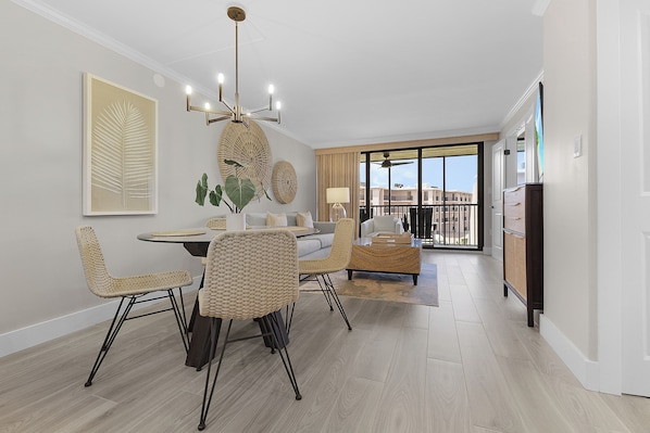 A modern living room with a dining area, featuring a chandelier, wicker furniture, and a large window with a view.