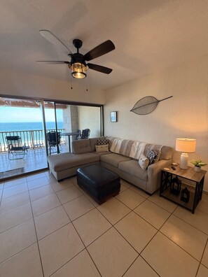 A living room with a sectional sofa, a ceiling fan, a view of the ocean, and a small table with a lamp.