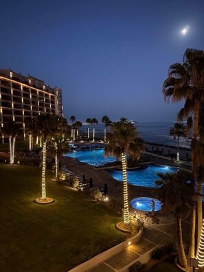 A hotel with a pool area, palm trees, and a view of the ocean at night.