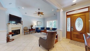 A spacious living room with a fireplace, a large TV, a blue sofa set, a ceiling fan, and a wooden door with a stained glass window.