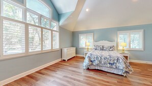 A bedroom with a large bed, two bedside tables with lamps, a dresser, and large windows with blinds.