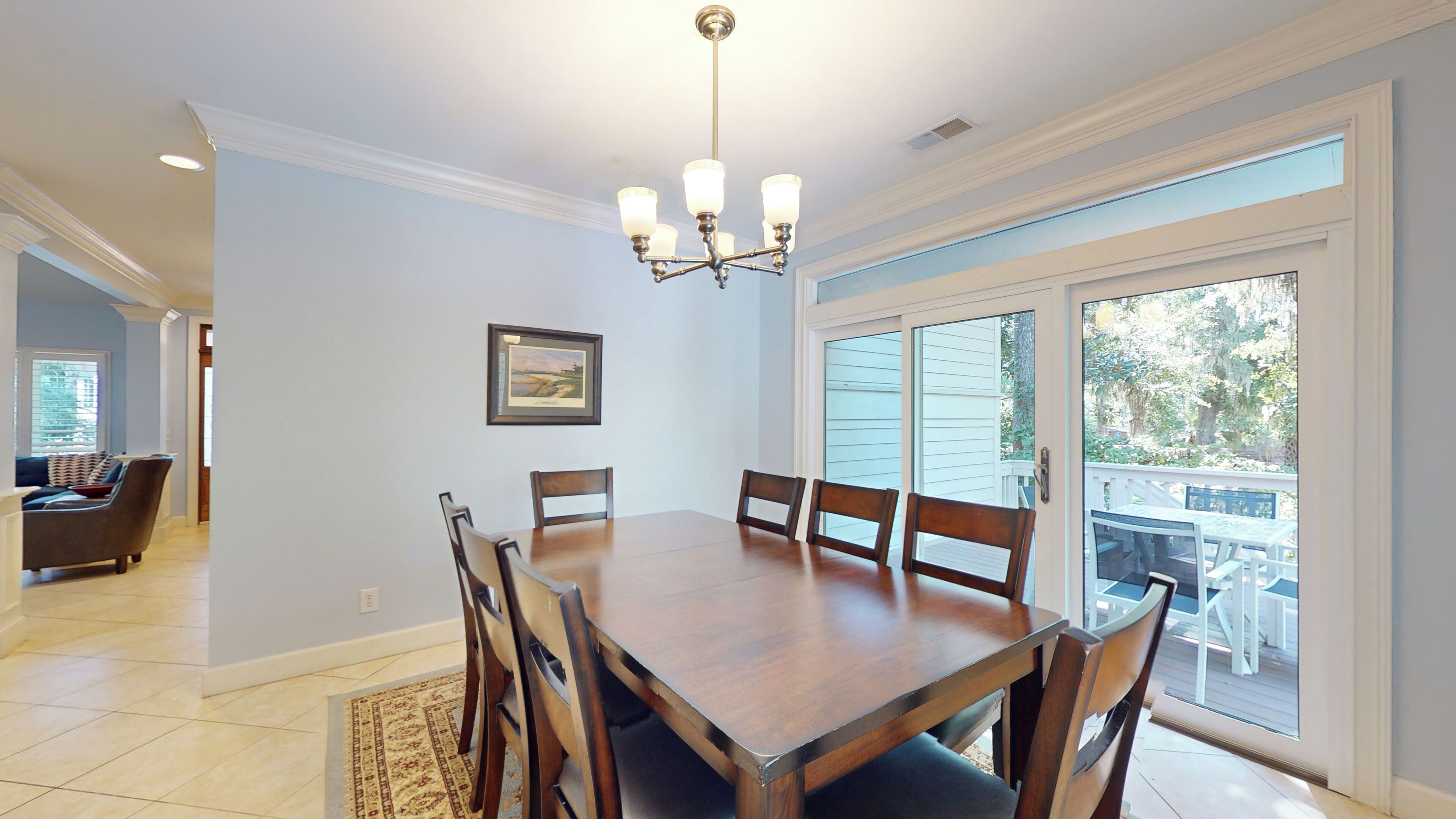 A dining room with a wooden table and chairs, a chandelier, a framed picture on the wall, and a view through a glass door to an outdoor deck.