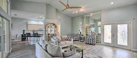 A spacious living room with a grey sofa, a coffee table, and a ceiling fan. There is a kitchen area in the background with modern appliances and a dining area with black chairs.
