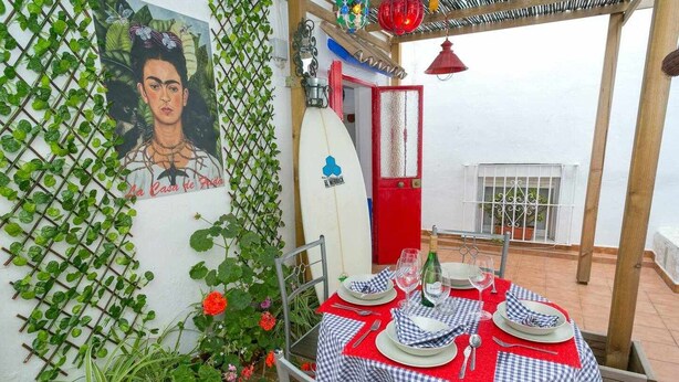 Una zona de menjador amb una taula preparada per a dues persones, una porta vermella i una paret amb un mural.