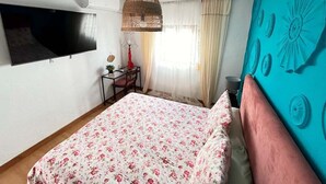 Un dormitori amb un llit, una televisió penjada a la paret, una taula de treball amb una cadira i una finestra amb cortines.