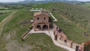Un bâtiment rustique en pierre, doté d’une cour, entouré de champs verdoyants et de collines lointaines.