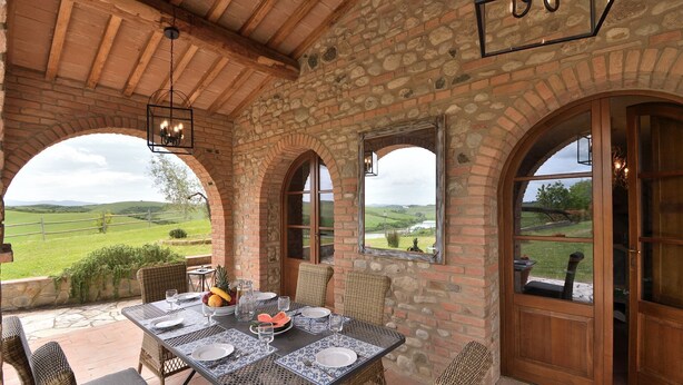 Une terrasse couverte avec une table dressée pour les repas, des murs en pierre et des fenêtres en arc donnant sur un paysage verdoyant.