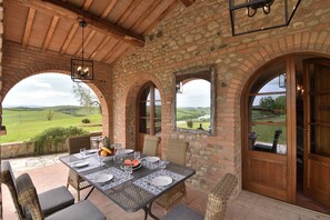Une terrasse couverte avec une table dressée pour les repas, des murs en pierre et des fenêtres en arc donnant sur un paysage verdoyant.