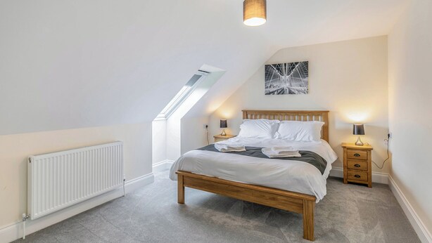 A bedroom with a sloped ceiling, a skylight, a bed with white bedding, two bedside tables with lamps, and a wall-mounted artwork.