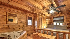 Interior de una cabaña de madera con una bañera de madera, una cama con cabecera, un ventilador de techo y una ventana con persianas.