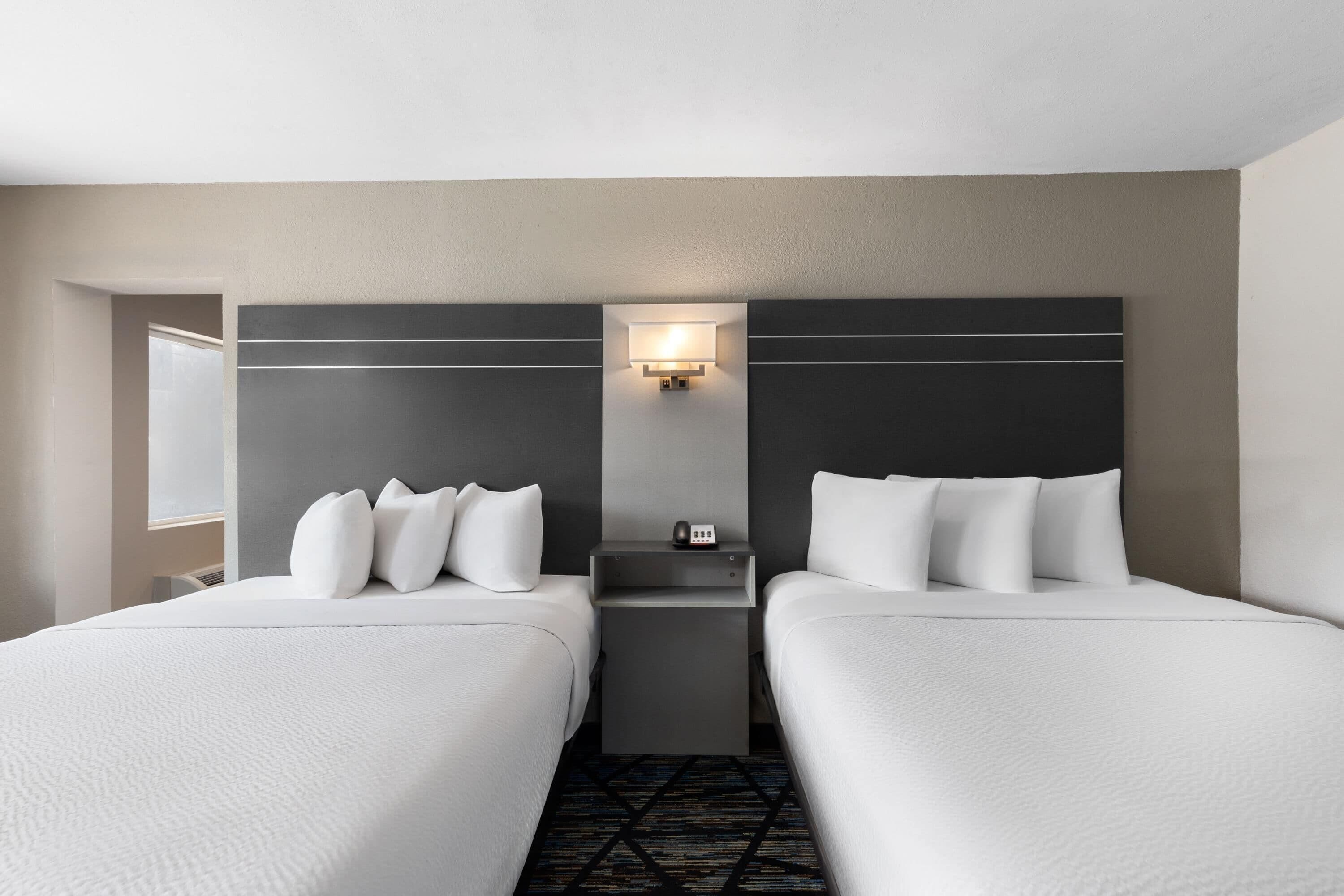 A hotel room with two beds, a nightstand, a wall-mounted lamp, and a window.